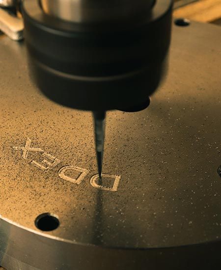Gros plan sur une machine gravant « DDEX » sur une surface métallique. Un foret est en action, au-dessus des lettres.