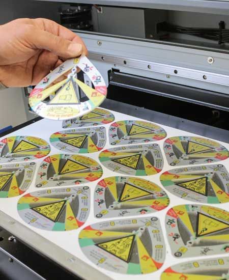 Une main tient une étiquette imprimée, détachée d'une feuille. Les étiquettes sont colorées et comportent un triangle d'avertissement au centre. L'image se trouve devant une machine d'impression.