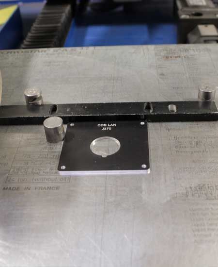 Carré noir avec une découpe circulaire sur une surface métallique, maintenu en place par des cylindres métalliques.