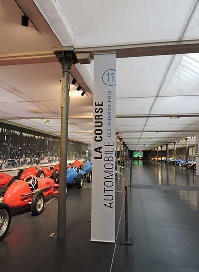 Des voitures de course sont exposées dans un musée. Un panneau indique « La Course Automobile ».