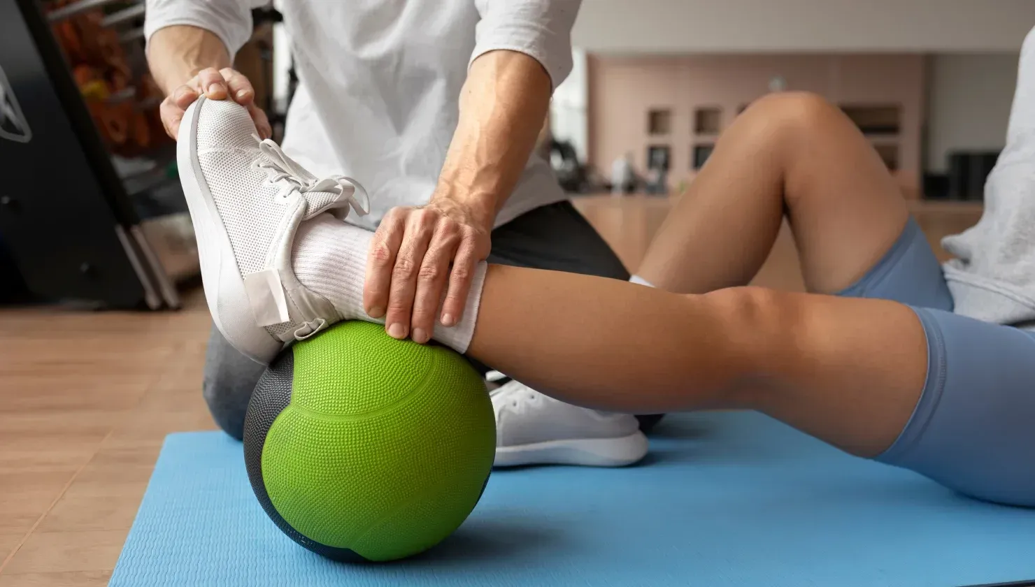 La gamba di una persona poggia su una palla verde, aiutata dalle mani di un'altra persona. Su un tappetino blu.