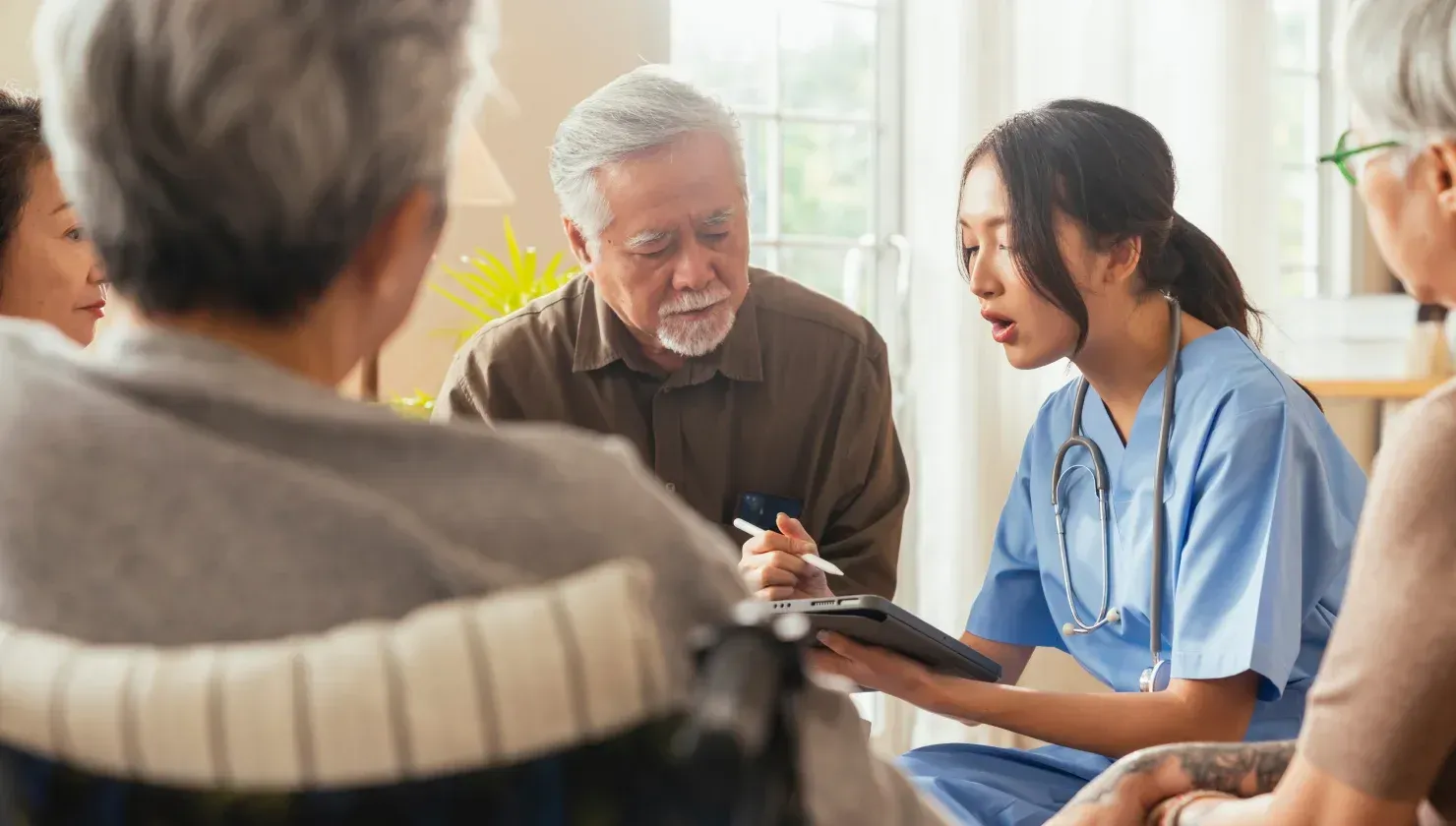 Un'infermiera in camice blu esamina il tablet insieme a un gruppo di anziani in una stanza.