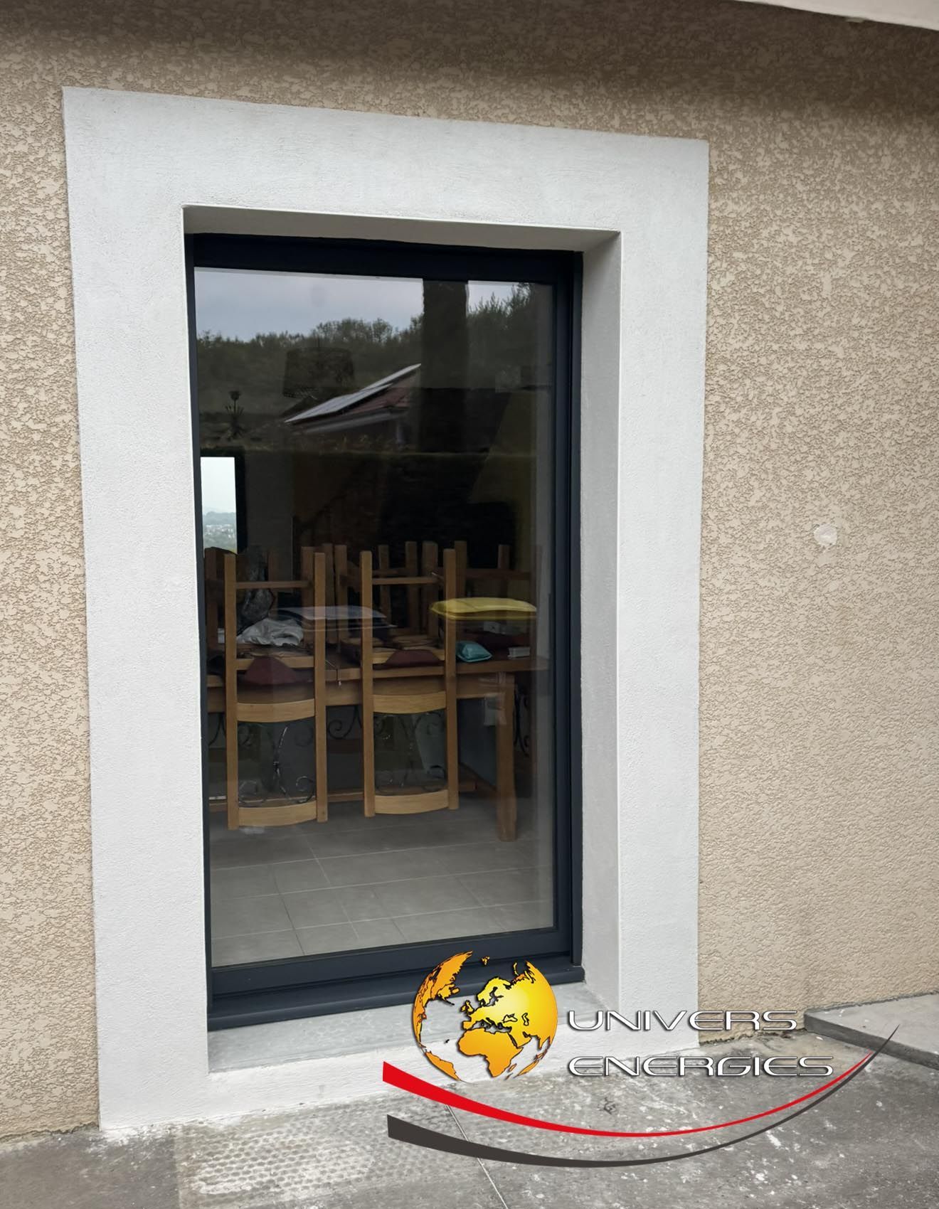 Fenêtre à cadre gris encastrée dans un mur en stuc beige, reflétant une scène de salle à manger intérieure.