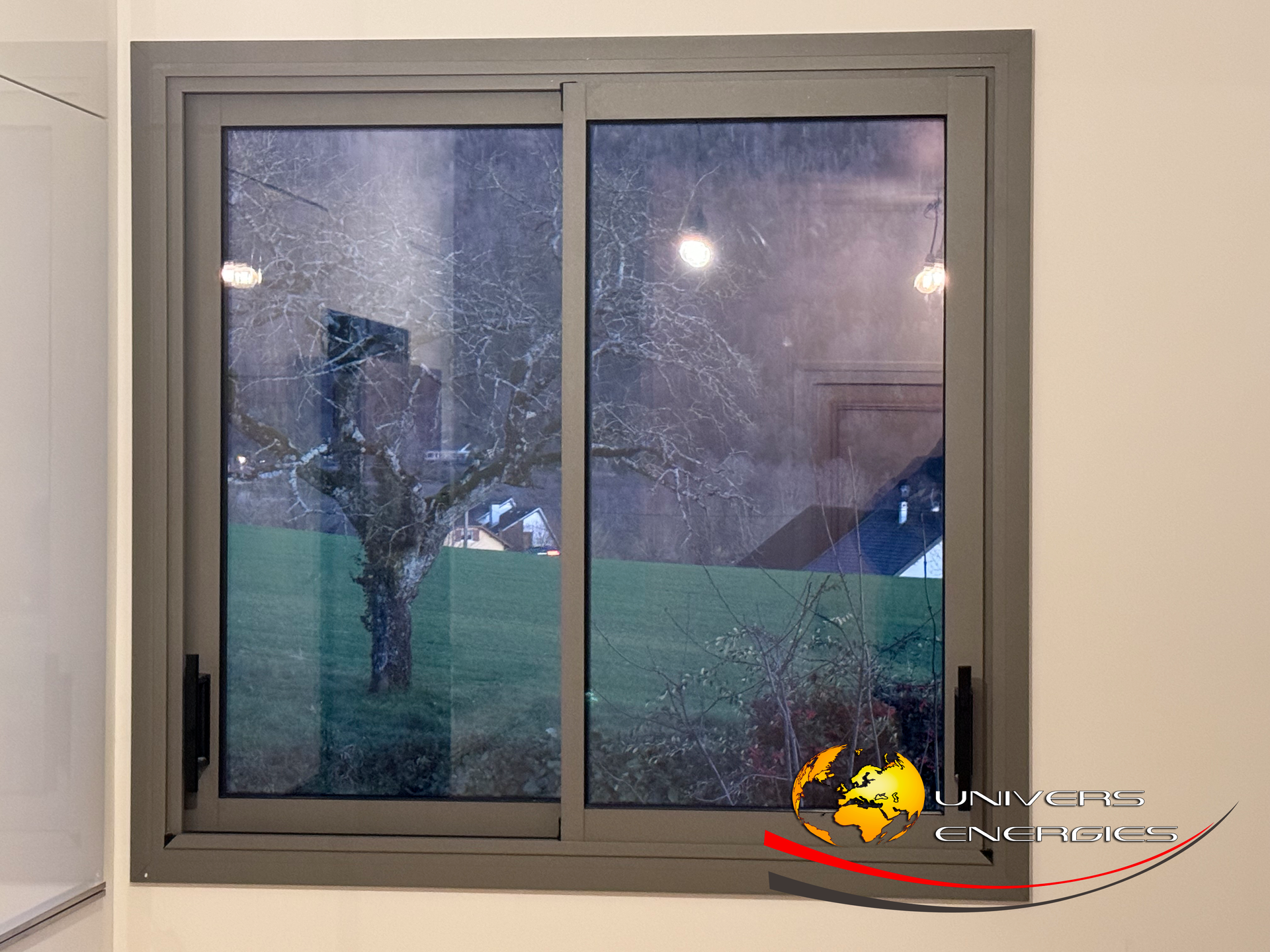 Fenêtre coulissante à cadre gris ; vue sur un arbre et un champ vert visibles à travers la vitre.