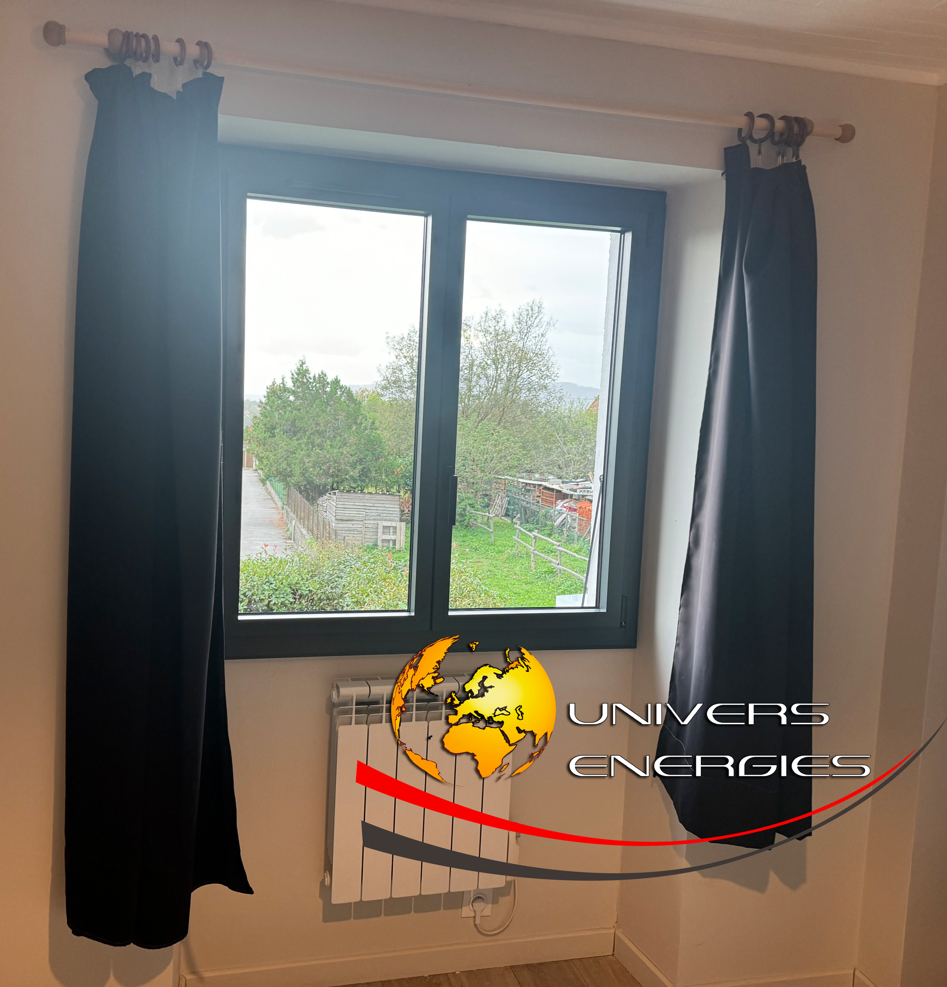 Fenêtre avec rideaux noirs et radiateur en dessous, donnant sur un espace extérieur verdoyant.