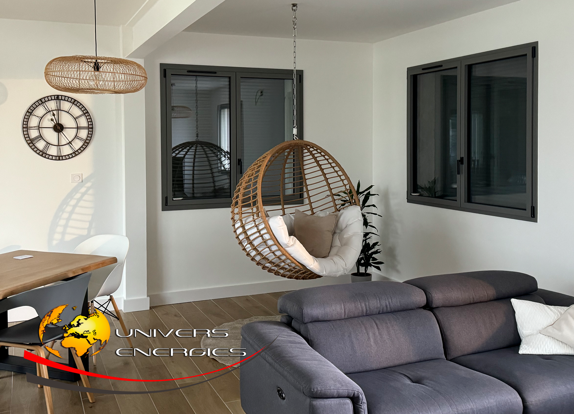 Salon avec canapé gris, fauteuil suspendu, fenêtres et horloge décorative.