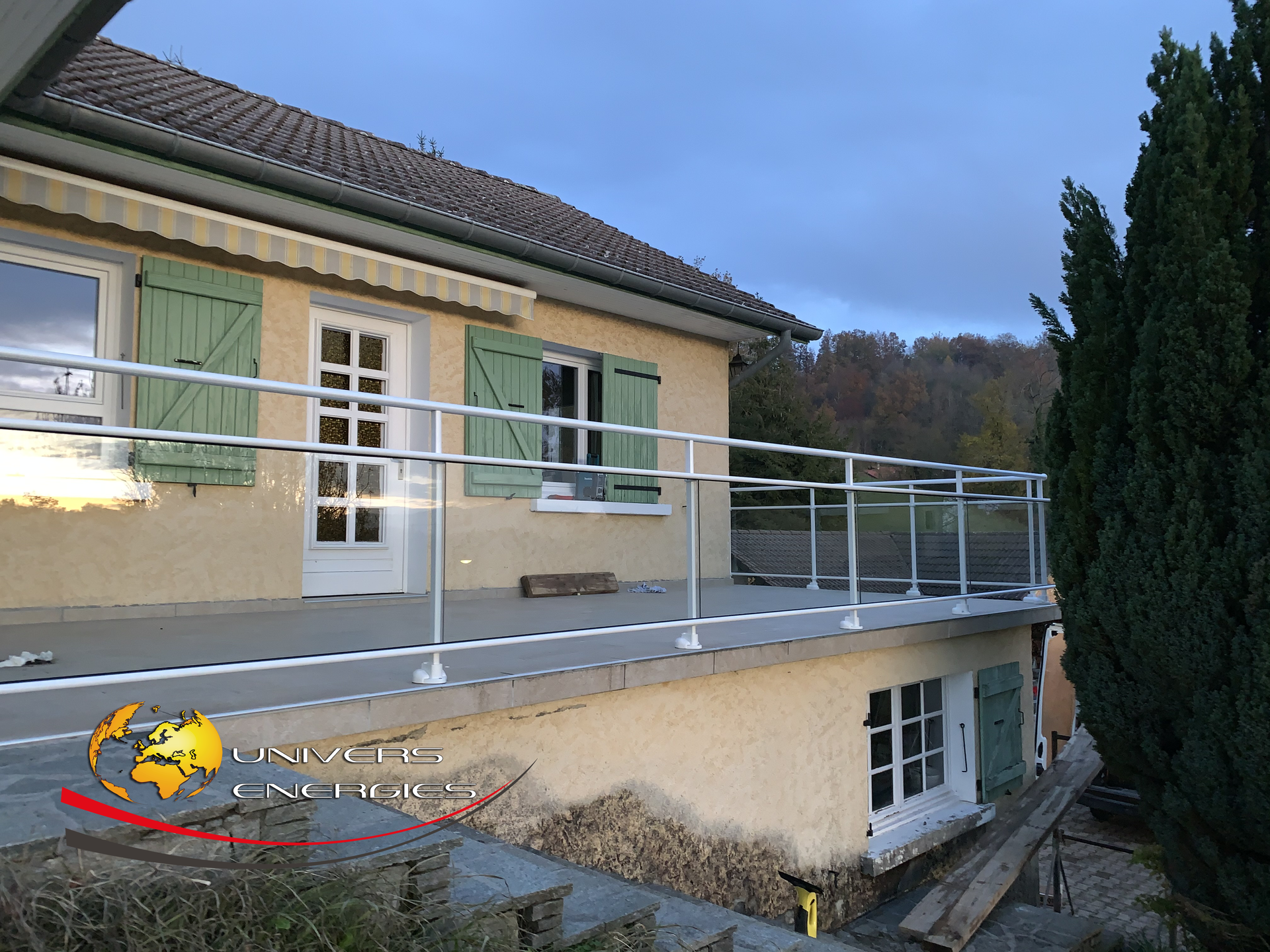 Vue extérieure d'une maison avec un balcon en béton doté de garde-corps en verre et en métal. Volets verts et ciel nuageux.