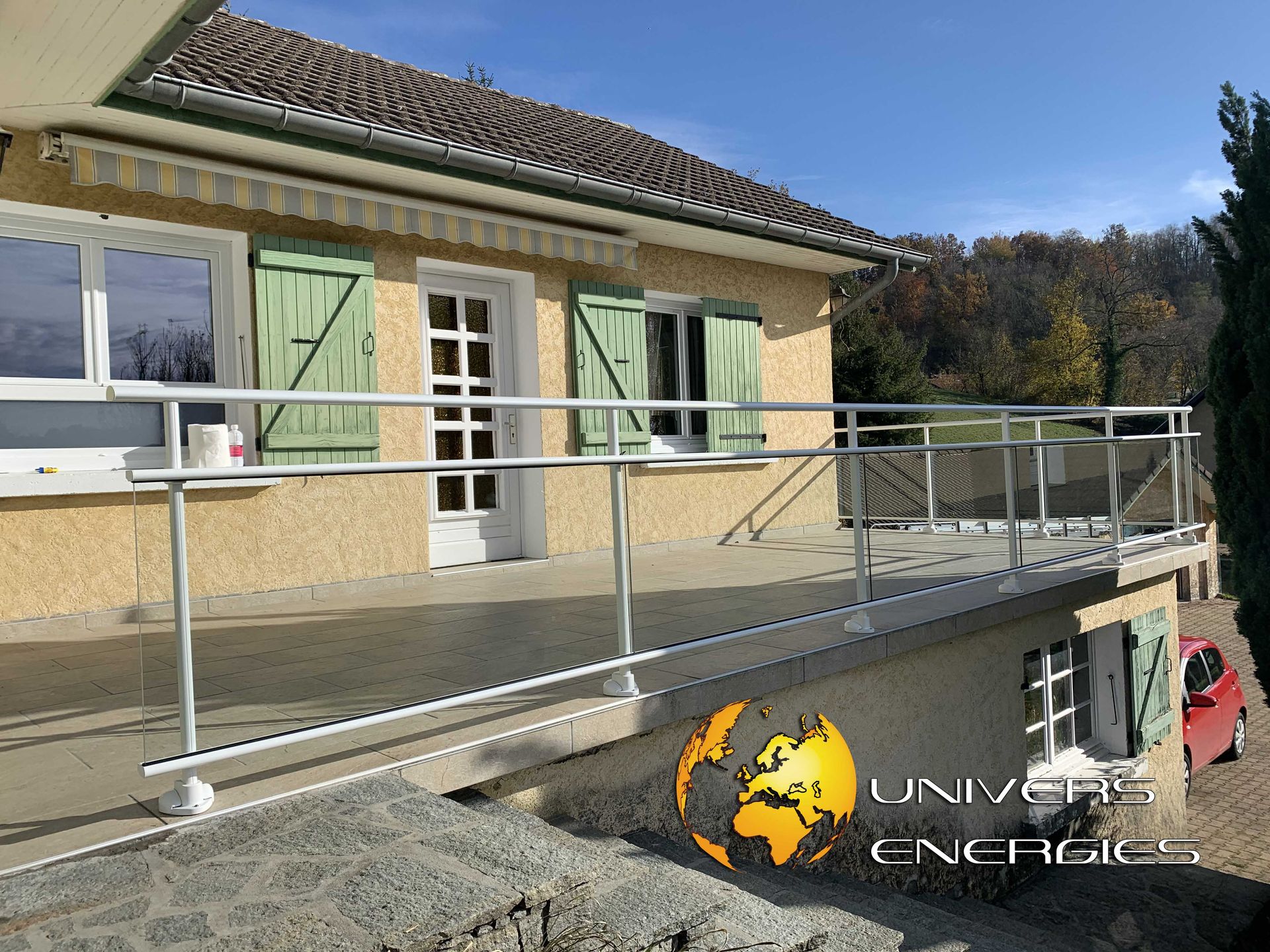 Rambarde en aluminium sur la terrasse d'une maison. Murs beiges, volets verts, extérieur ensoleillé.
