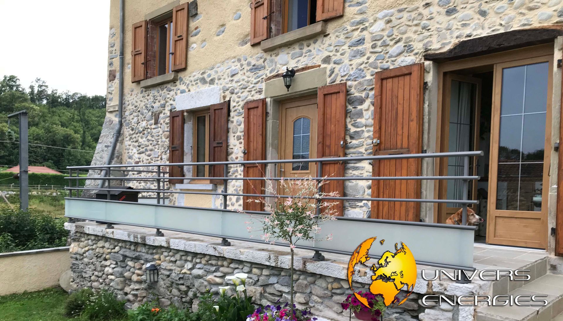 Maison en pierre avec volets bruns, balustrade en verre et porte en bois ouverte.