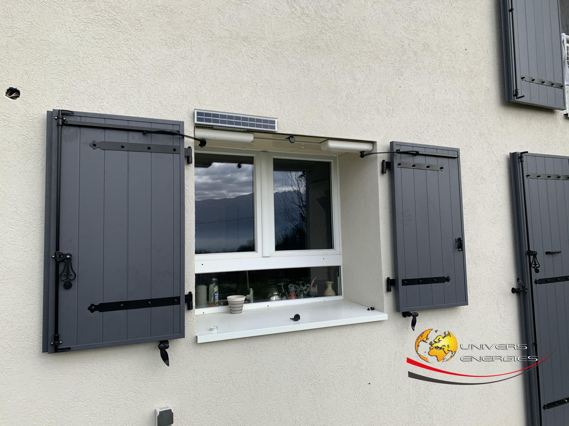 Des volets gris s'ouvrent à côté d'une fenêtre à cadre blanc sur un bâtiment clair.