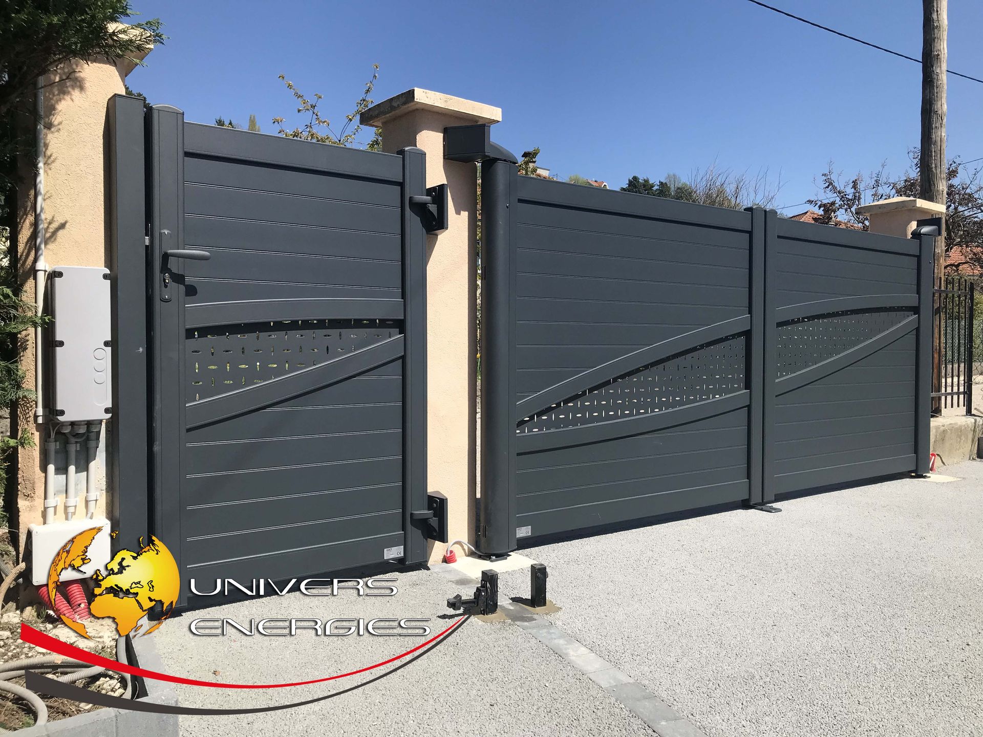 Portail en métal gris foncé à motif diagonal, allée de gravier, colonnes beiges, ciel bleu.
