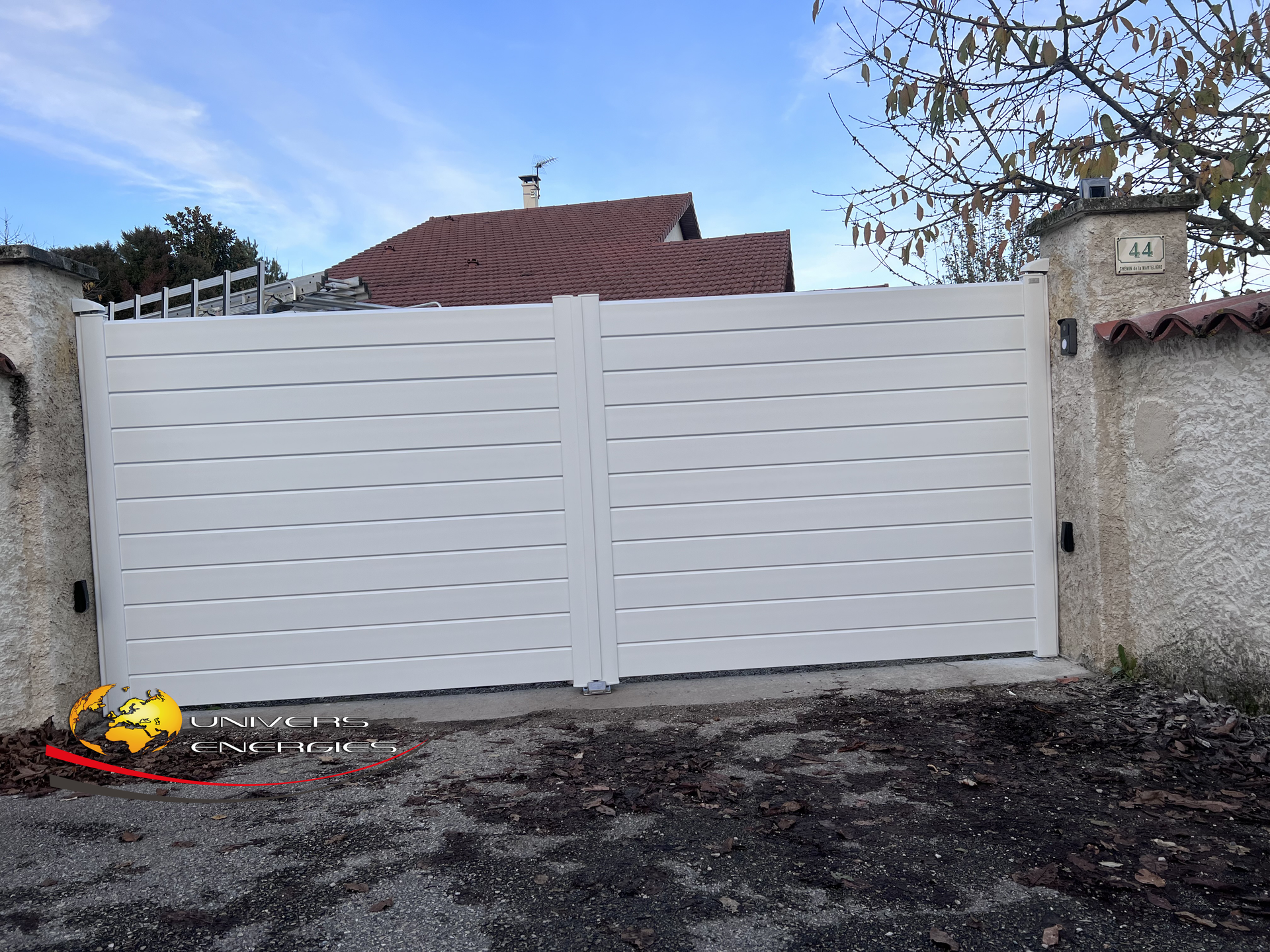 Portail gris clair à lames horizontales, fermé, devant une maison, placé entre deux piliers de pierre.