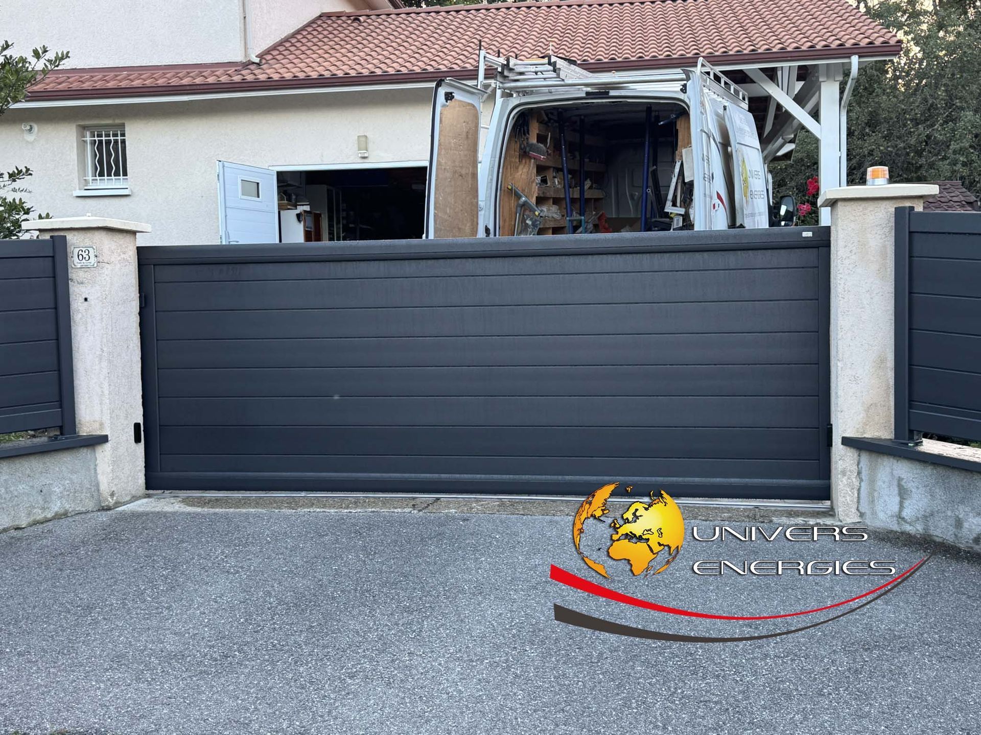Portail coulissant gris foncé devant une maison. Allée en granit avec un logo en bas.