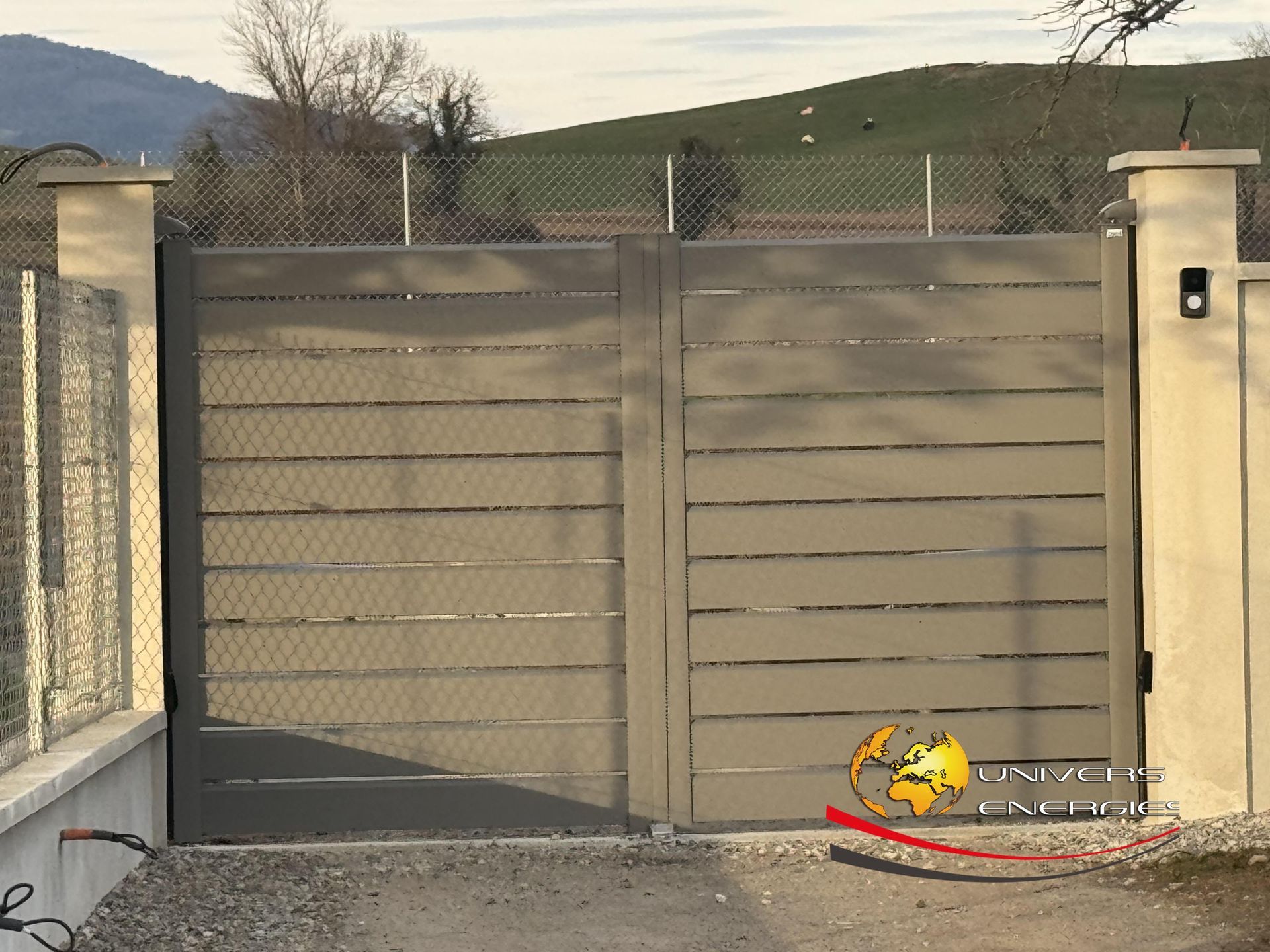 Portail en métal gris à lattes horizontales, allée de gravier, piliers en béton et clôture en grillage à l'arrière-plan.