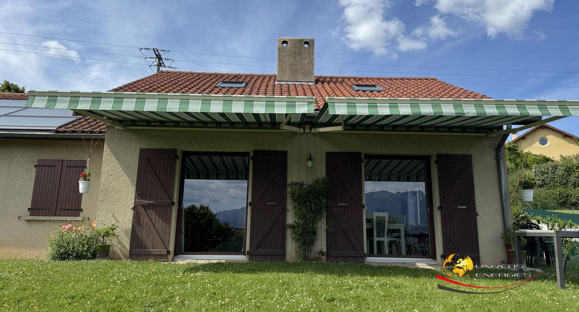 Maison en stuc beige avec auvent à rayures vertes, volets marron et cheminée se détachant sur un ciel bleu.