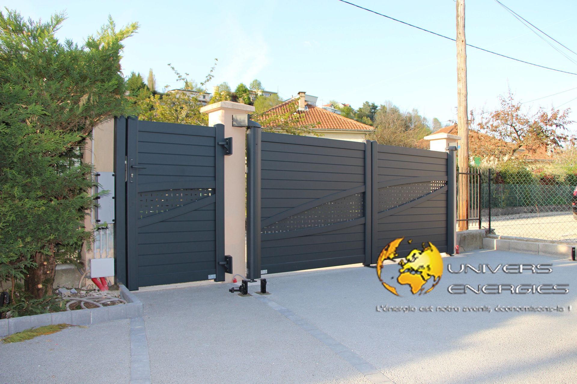 Un portail métallique gris s'ouvre sur une allée carrossable, flanquée de piliers, avec des arbres et une maison en arrière-plan.