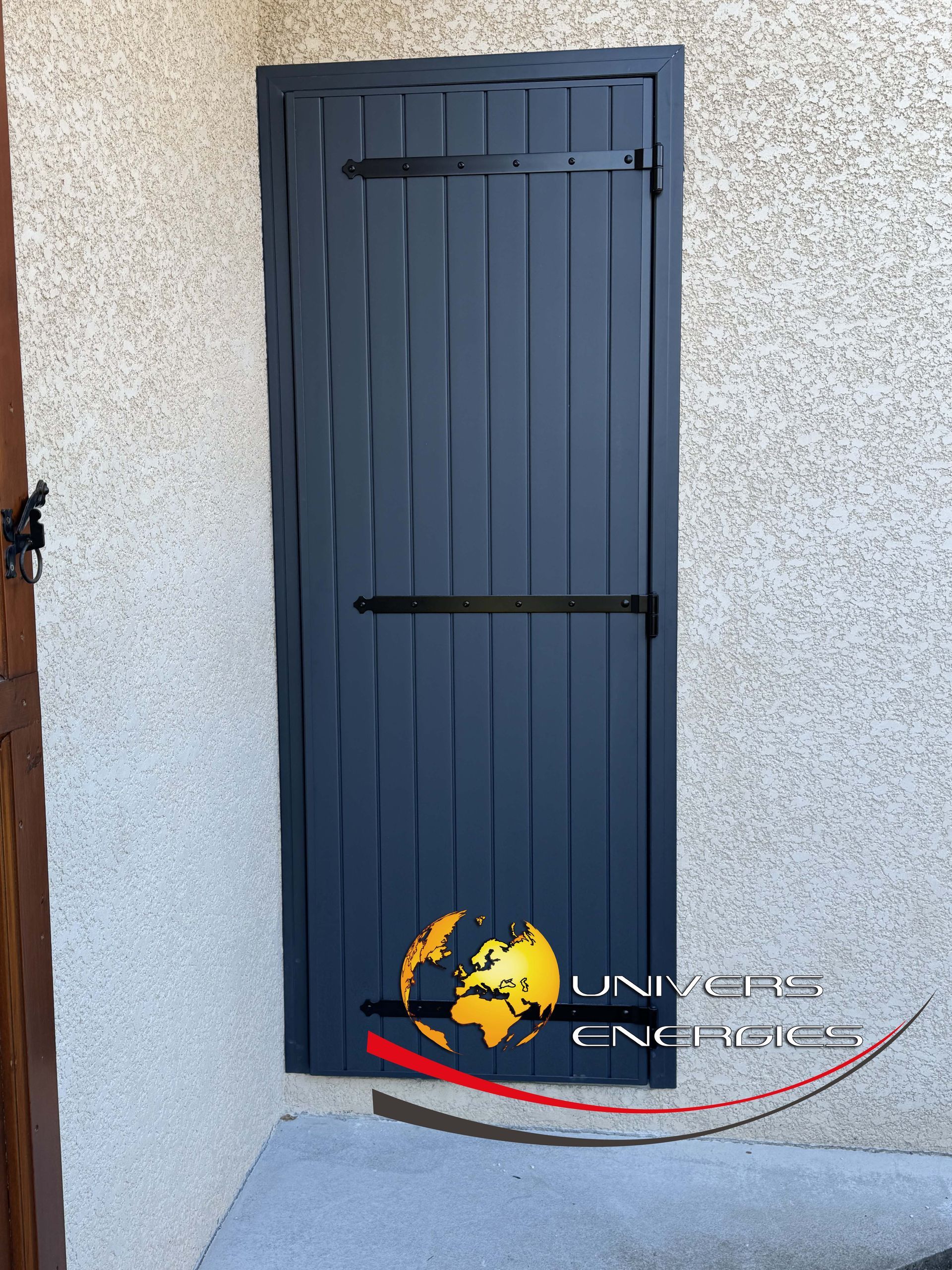 Porte gris foncé à rayures verticales, charnières et renforts noirs, encastrée dans un mur beige clair.