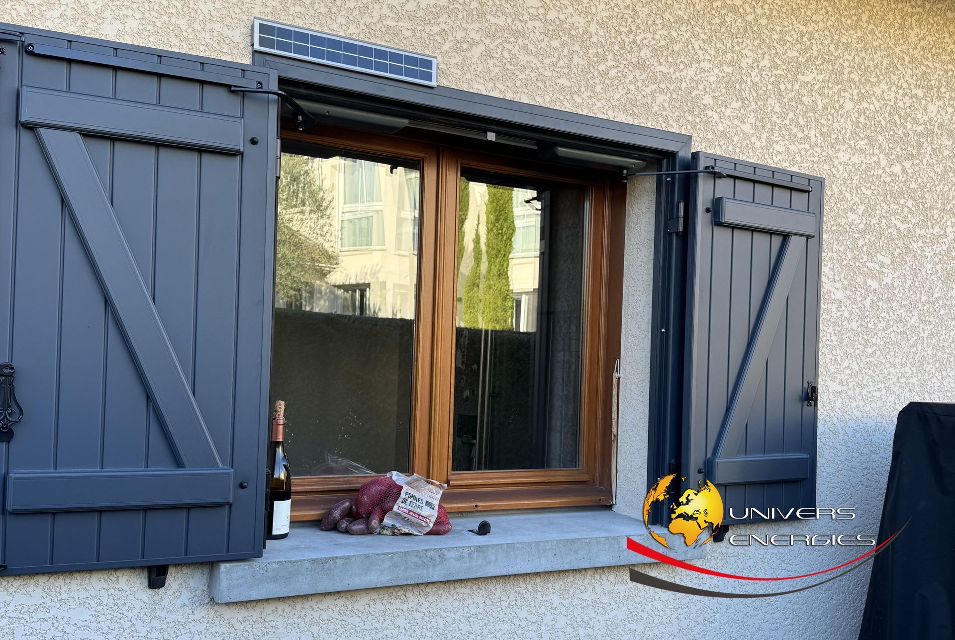 Fenêtre avec volets gris, cadre marron et petit panneau solaire. Vue extérieure.