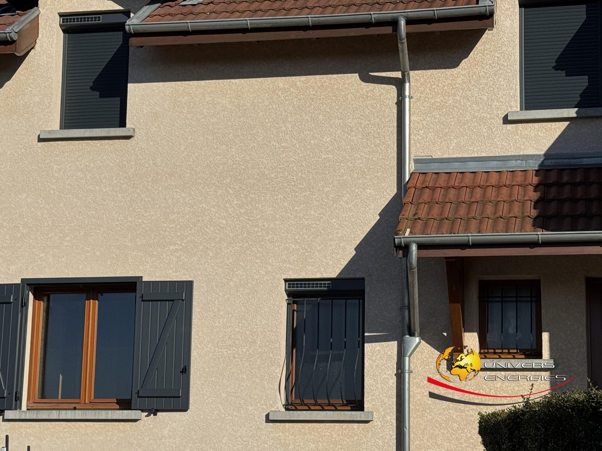 Bâtiment beige avec volets et fenêtres foncés ; une descente de gouttière longe le mur ; journée ensoleillée.