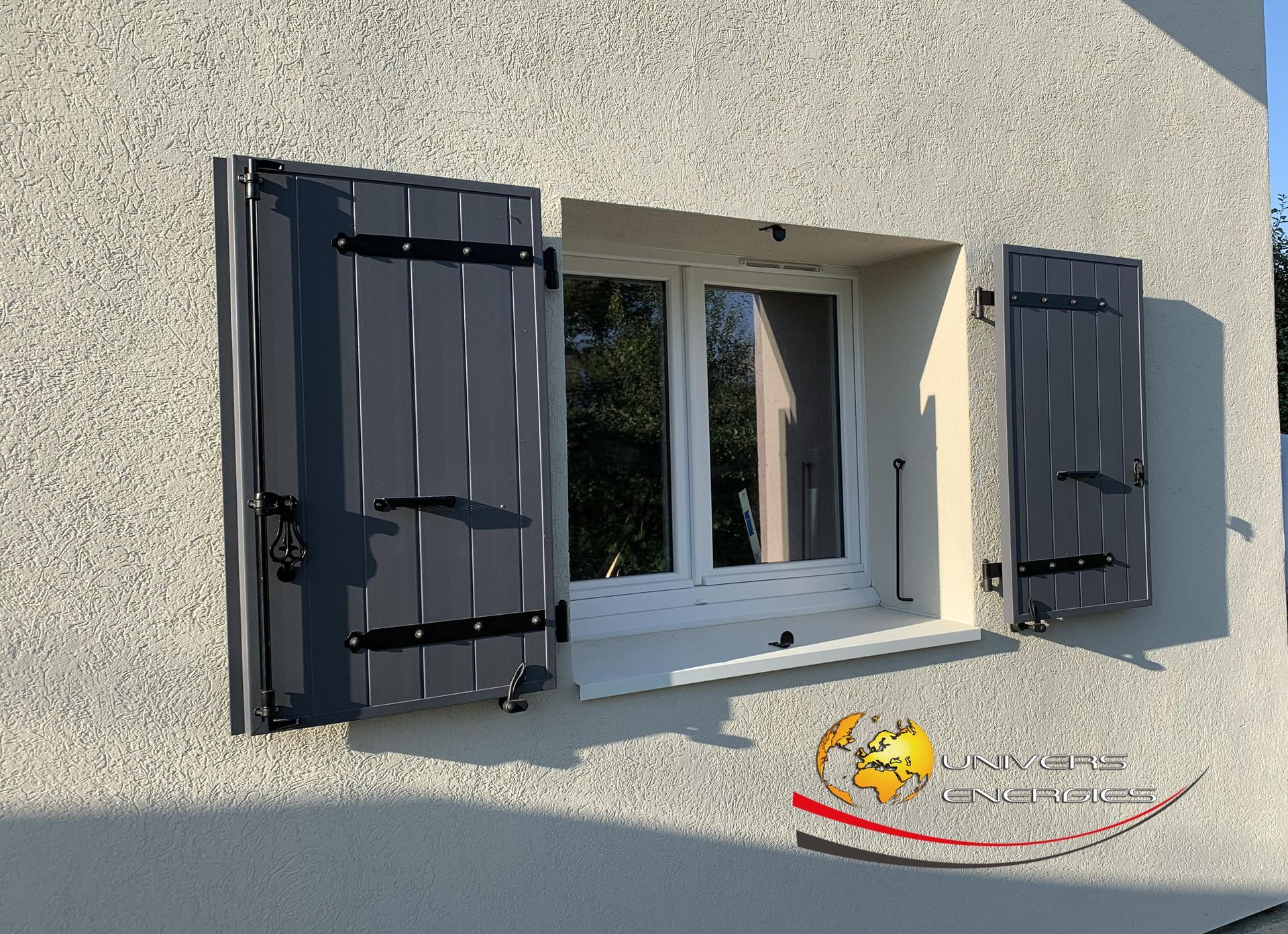 Fenêtre à volets gris sur un mur en stuc. Les volets sont ouverts, laissant apparaître la fenêtre.