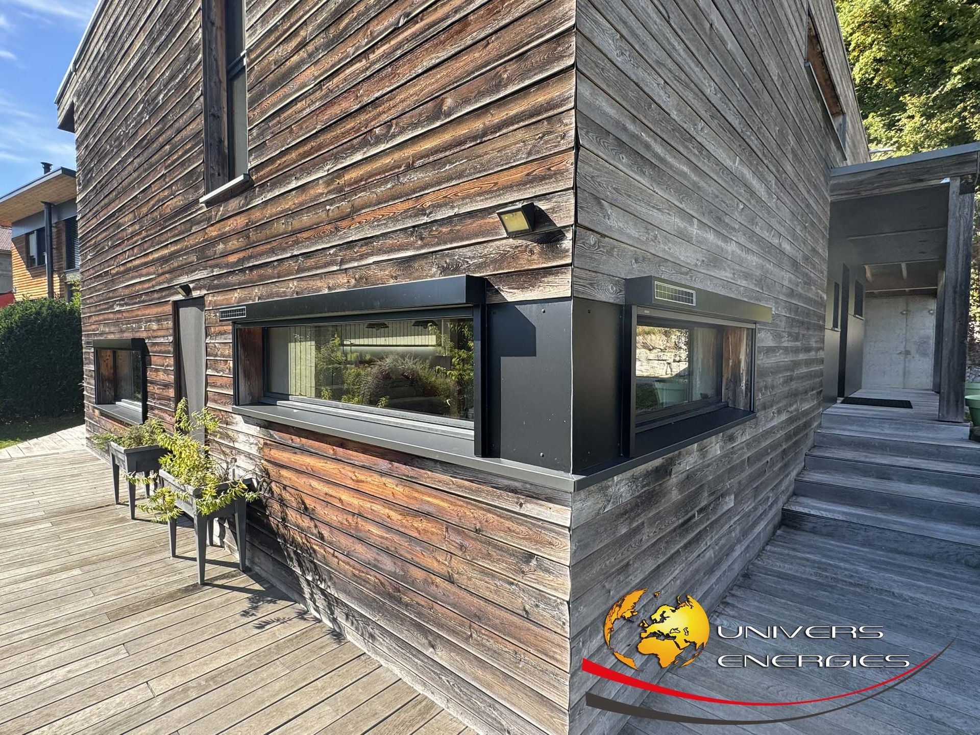 Bâtiment en bois aux fenêtres à cadre noir ; vue extérieure. Une terrasse et des marches en bois mènent à une entrée.