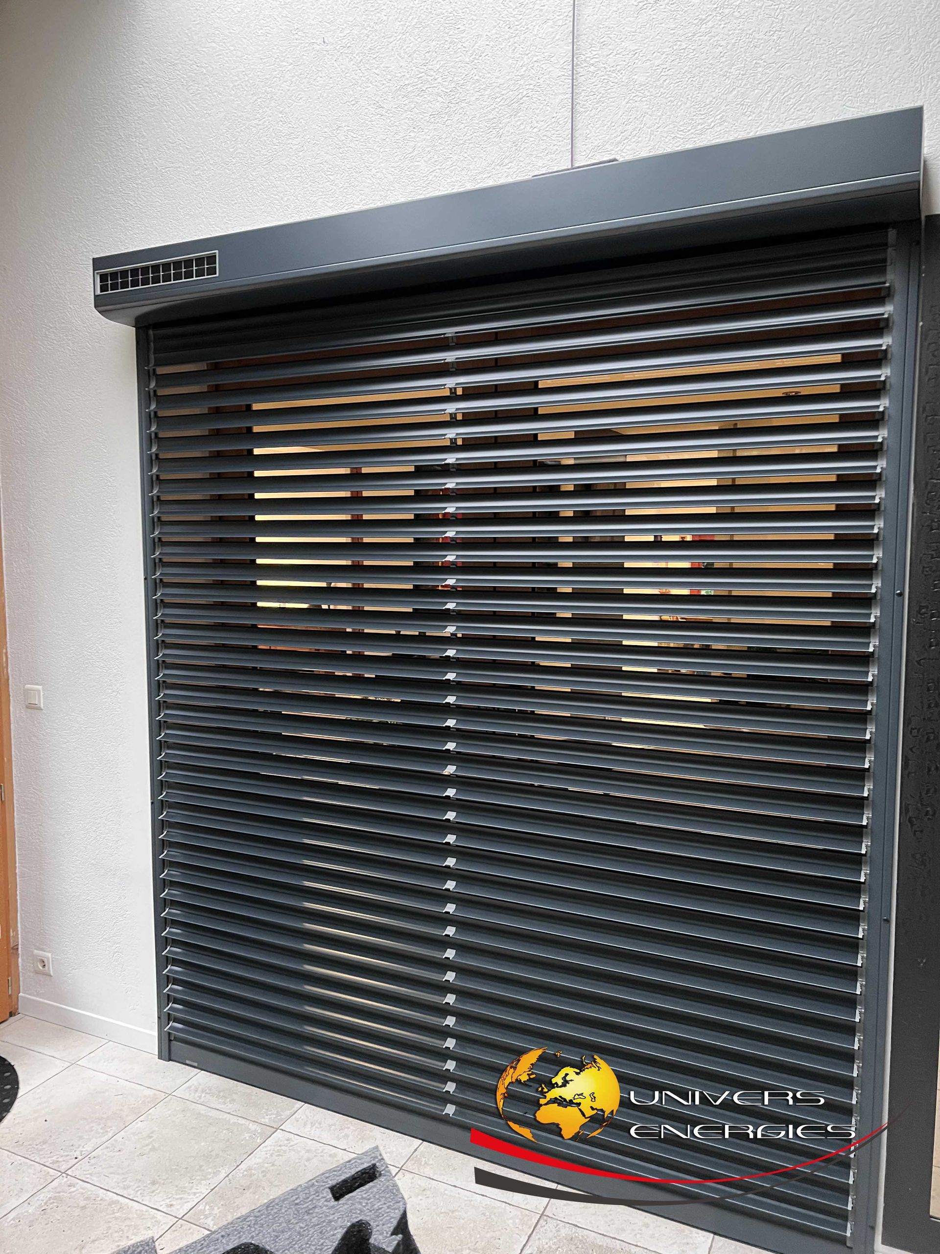 Volet de sécurité métallique gris foncé recouvrant une fenêtre, fixé sur un mur gris clair.