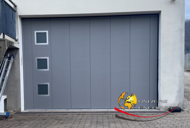 Porte de garage grise avec trois fenêtres carrées, logo et câble rouge sur béton.