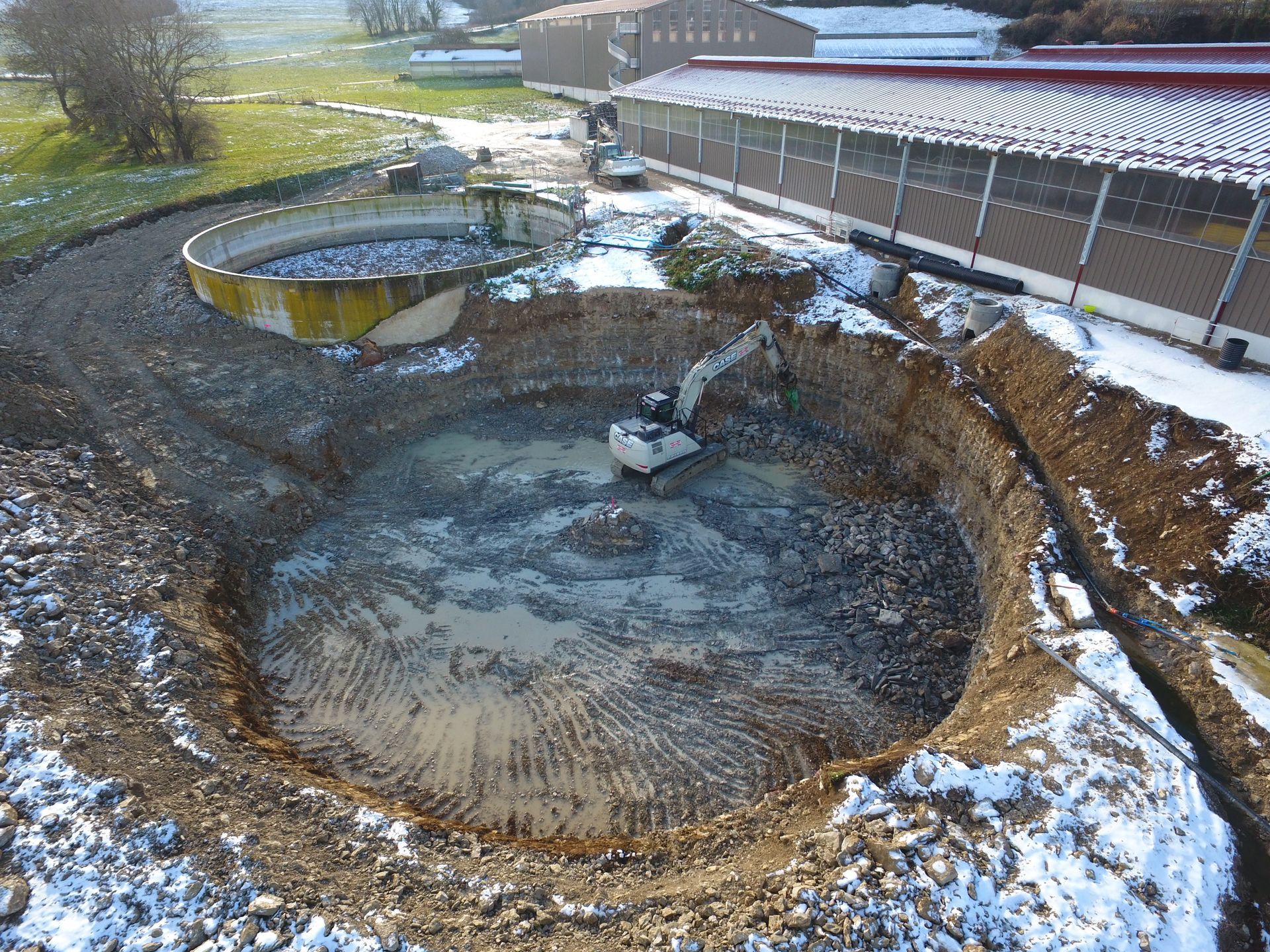 Excavatrice travaillant à l'intérieur d'une grande fosse circulaire près d'un bâtiment agricole ; sol enneigé.