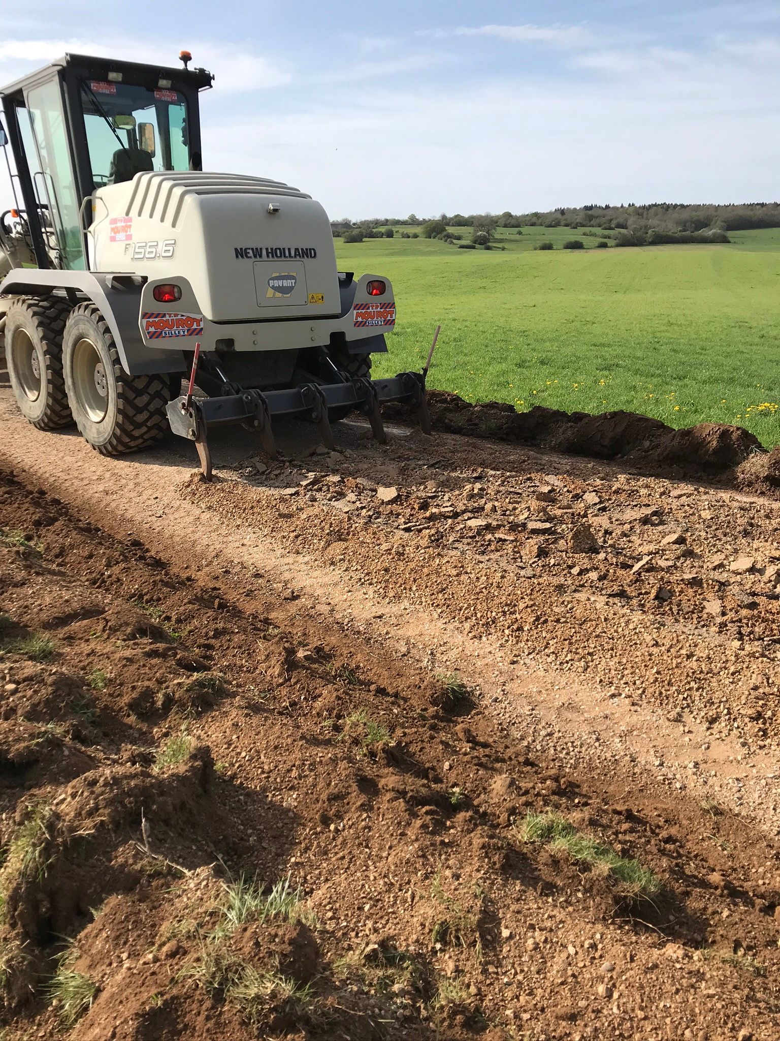 Une niveleuse, gris clair, nivelant un chemin de terre à côté d'un champ vert sous un ciel bleu.