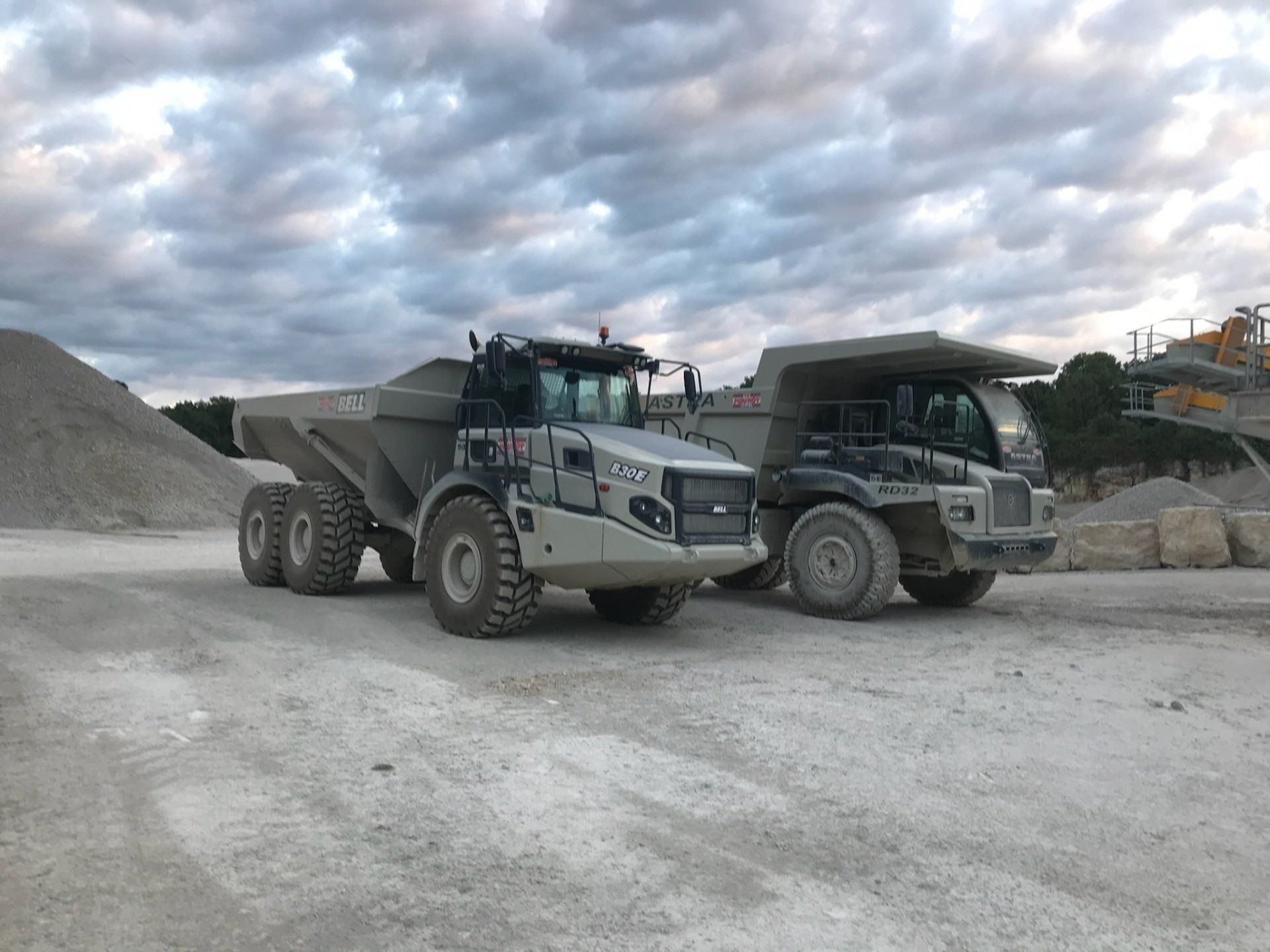 Deux camions à benne basculante gris garés sur une surface de gravier ; ciel nuageux au-dessus.