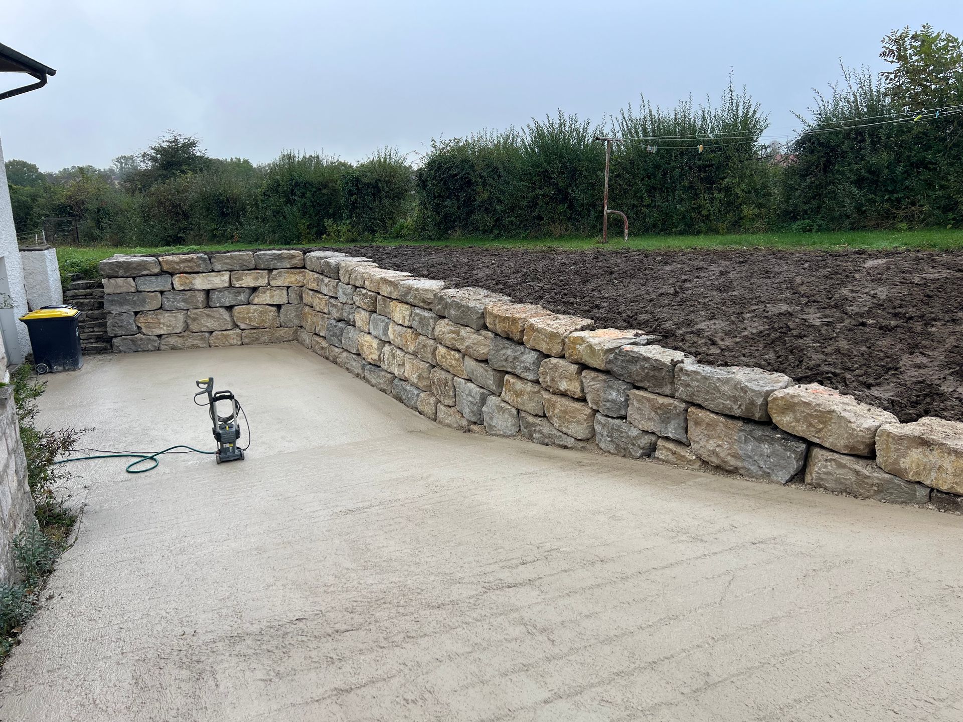 Un mur de soutènement en pierre borde un patio en béton et un jardin en terre.