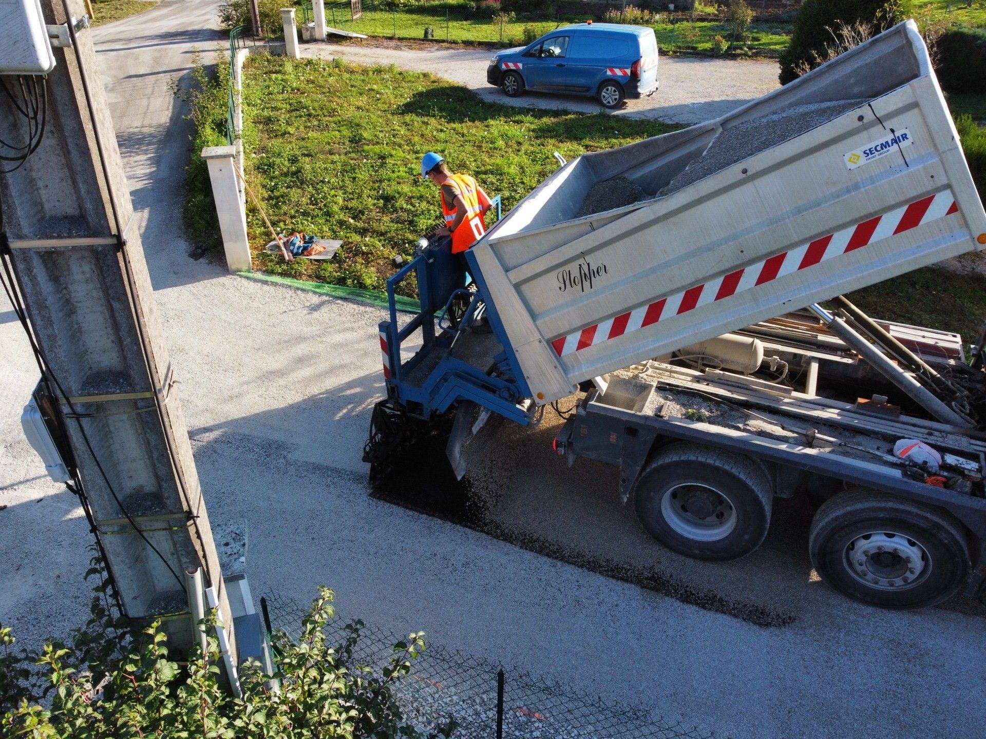Un ouvrier du bâtiment portant un gilet orange conduit un camion à benne basculante et dépose du gravier sur une allée.