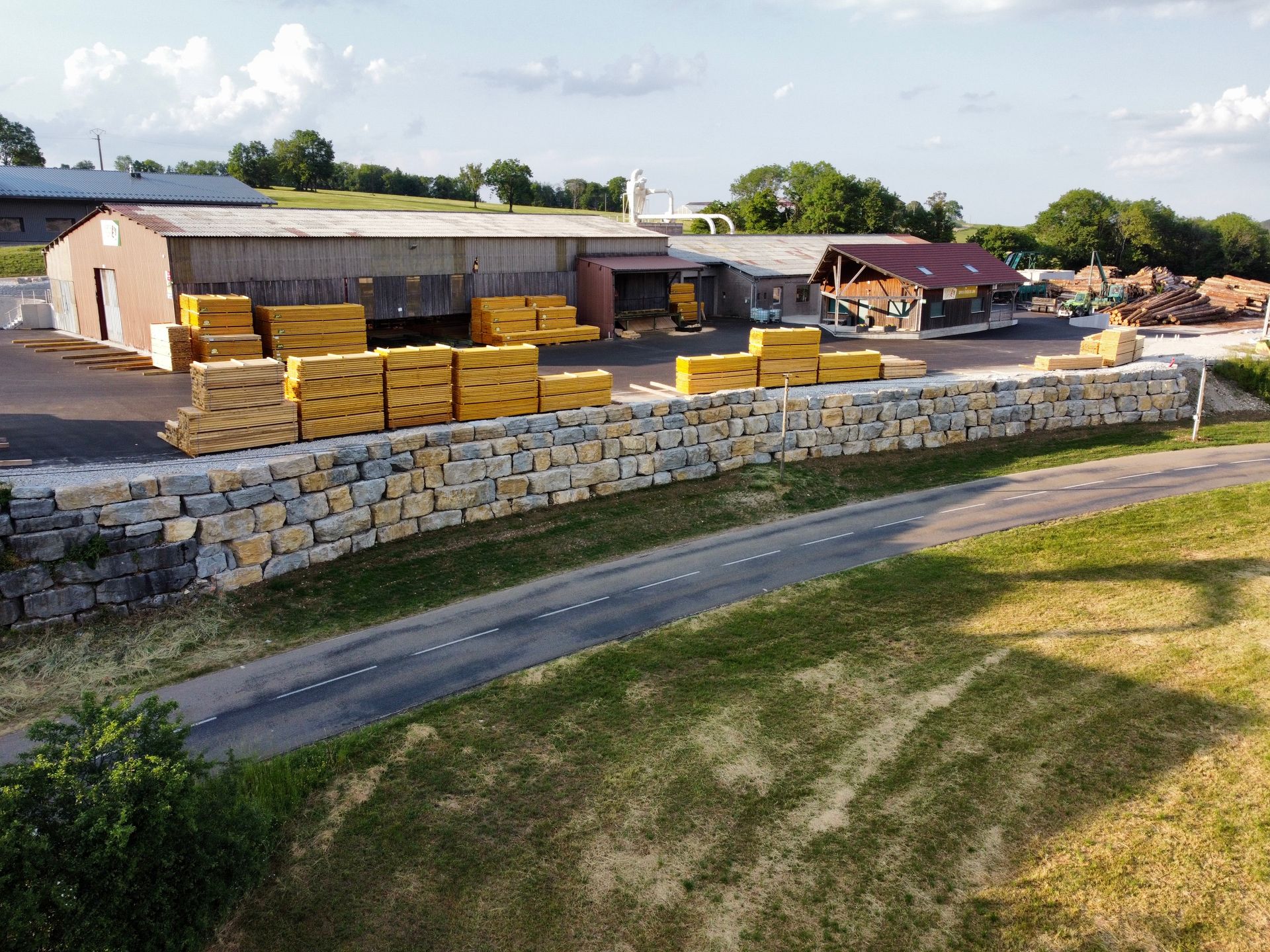Une cour à bois avec du bois jaune empilé, un mur de soutènement en pierre, des bâtiments et une route par une journée ensoleillée.