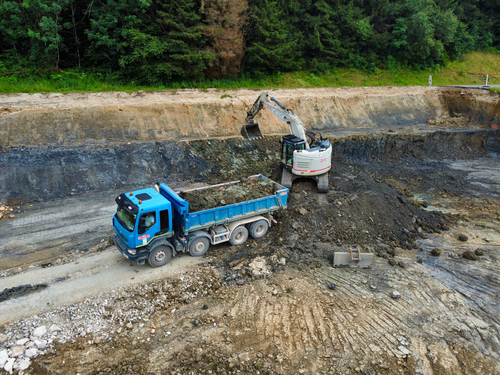Un camion à benne bleu chargé par une excavatrice sur un chantier de construction près d'une forêt.