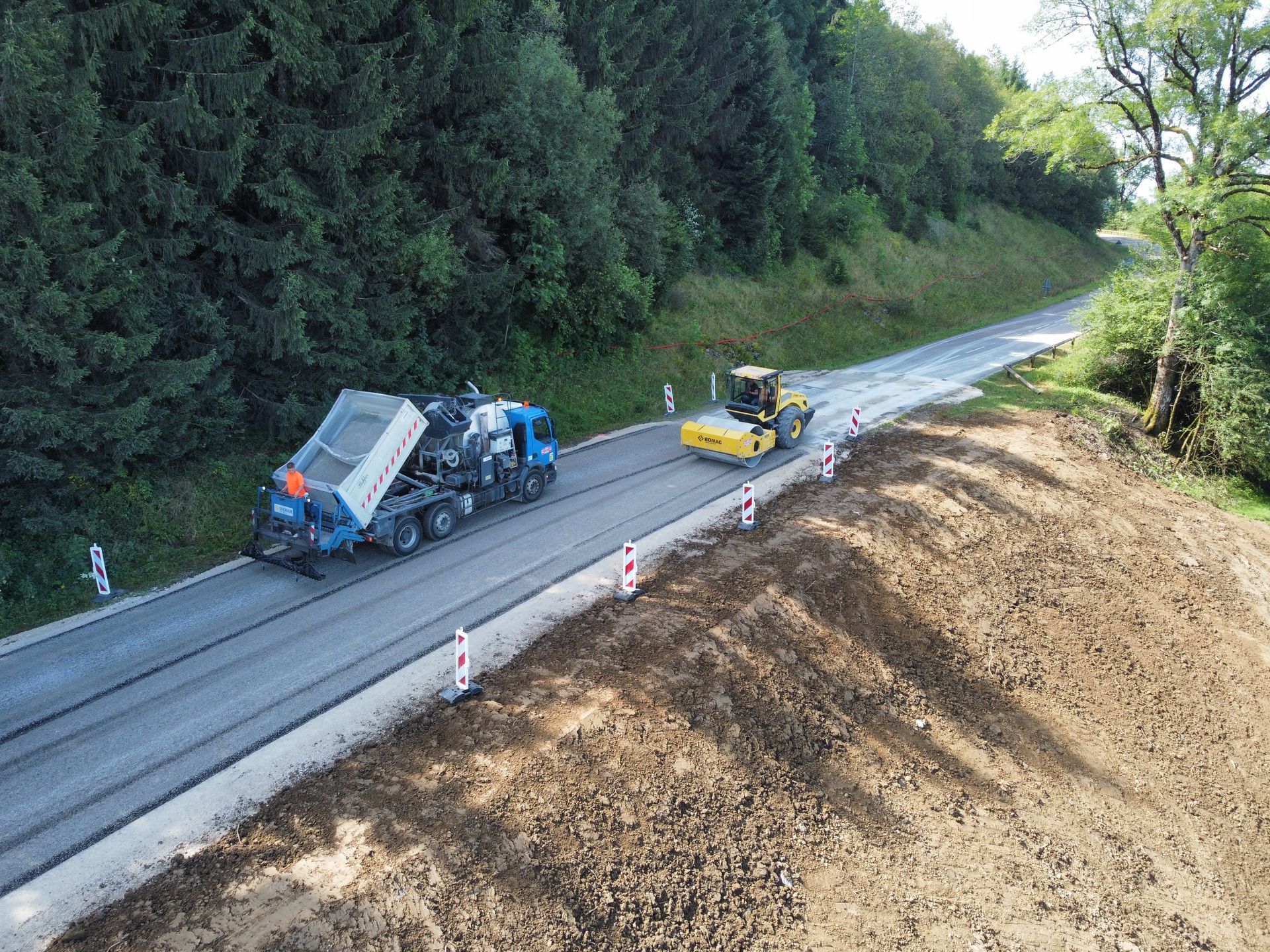 Travaux de pavage de route en cours ; une machine pose de l'asphalte, un rouleau le compacte, le long d'une colline boisée.