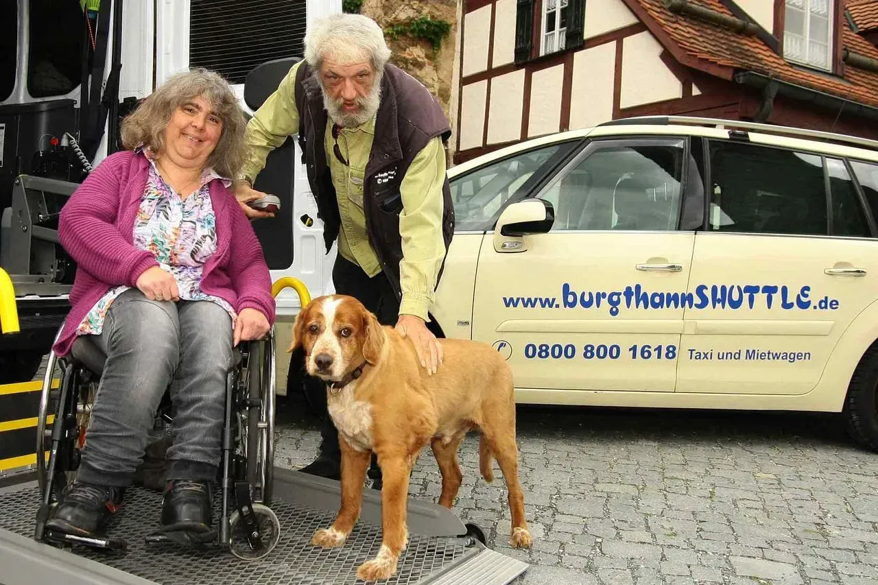 Rollstuhlfahrerin mit Fahrer und Hund vor einem Auto