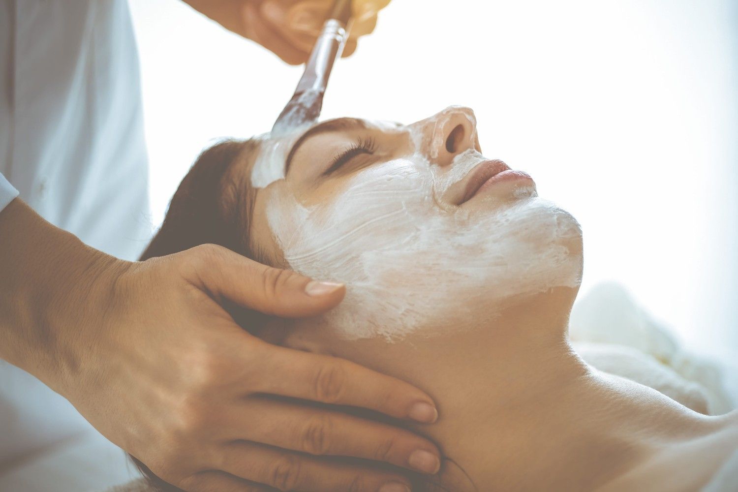 Tratamiento facial en curso: una mujer recibe una mascarilla de crema blanca y un masaje facial.