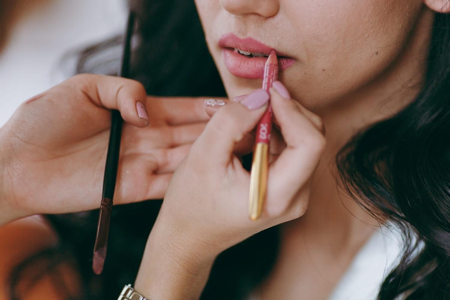 Una maquilladora aplica delineador de labios rosa en los labios de una mujer mientras sostiene un pincel para labios.