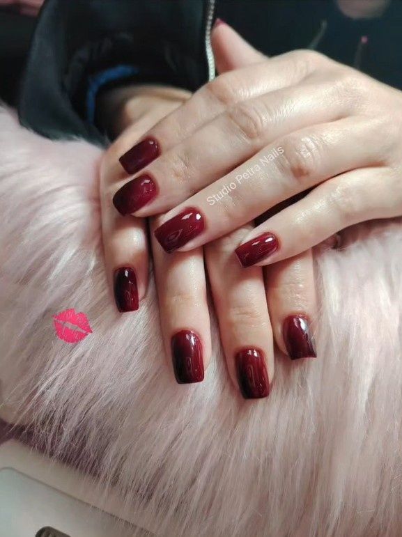 Manos bien cuidadas con uñas de color rojo oscuro descansando sobre una suave piel sintética rosa, con una pequeña marca de labios rosa cerca.