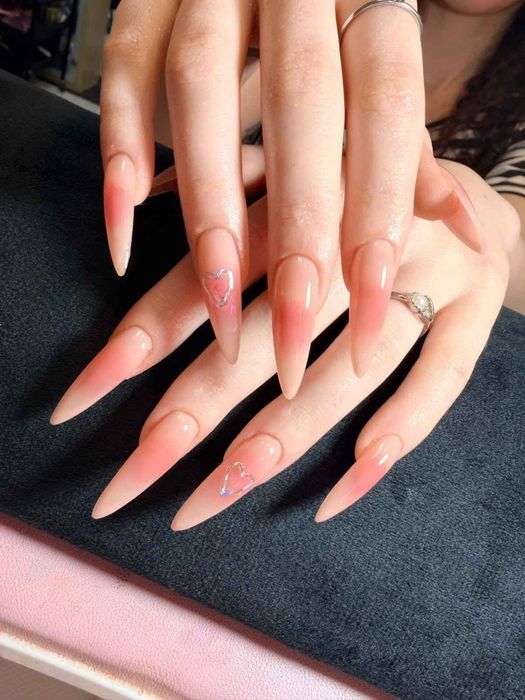 Uñas largas de color rosa degradado en forma de aguja sobre manos superpuestas con un anillo plateado, descansando sobre una superficie oscura.