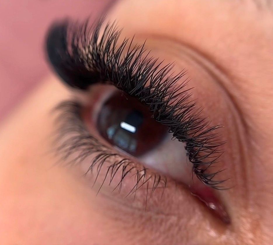 Primer plano de un ojo marrón con largas pestañas postizas rizadas y maquillaje rosa suave.