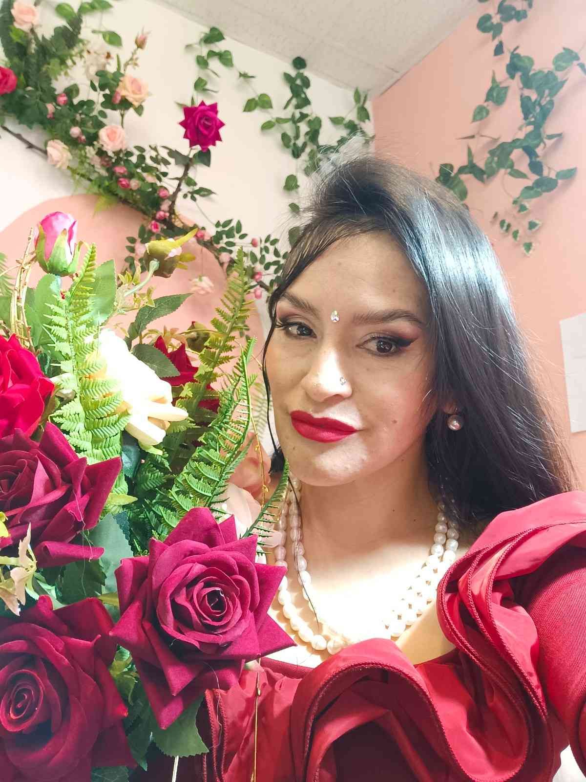 Mujer con un vestido rojo posando con rosas en una habitación decorada con flores, luciendo joyas de perlas.