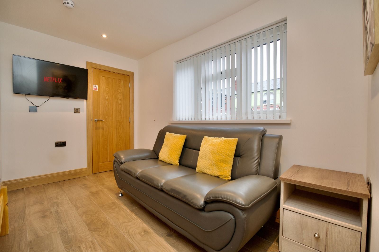 Small living room with gray sofa, yellow cushions, wall TV, wooden door, and window blinds