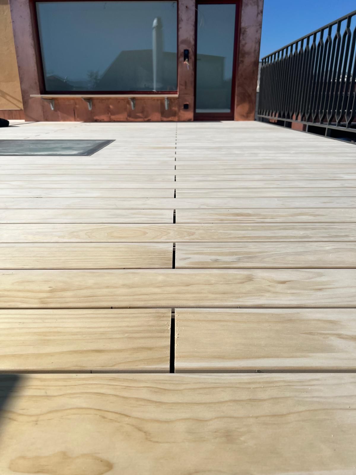 Holzdeck mit Lücken, das zu einem Gebäude mit Fenstern, einem schwarzen Geländer und einem blauen Himmel führt.