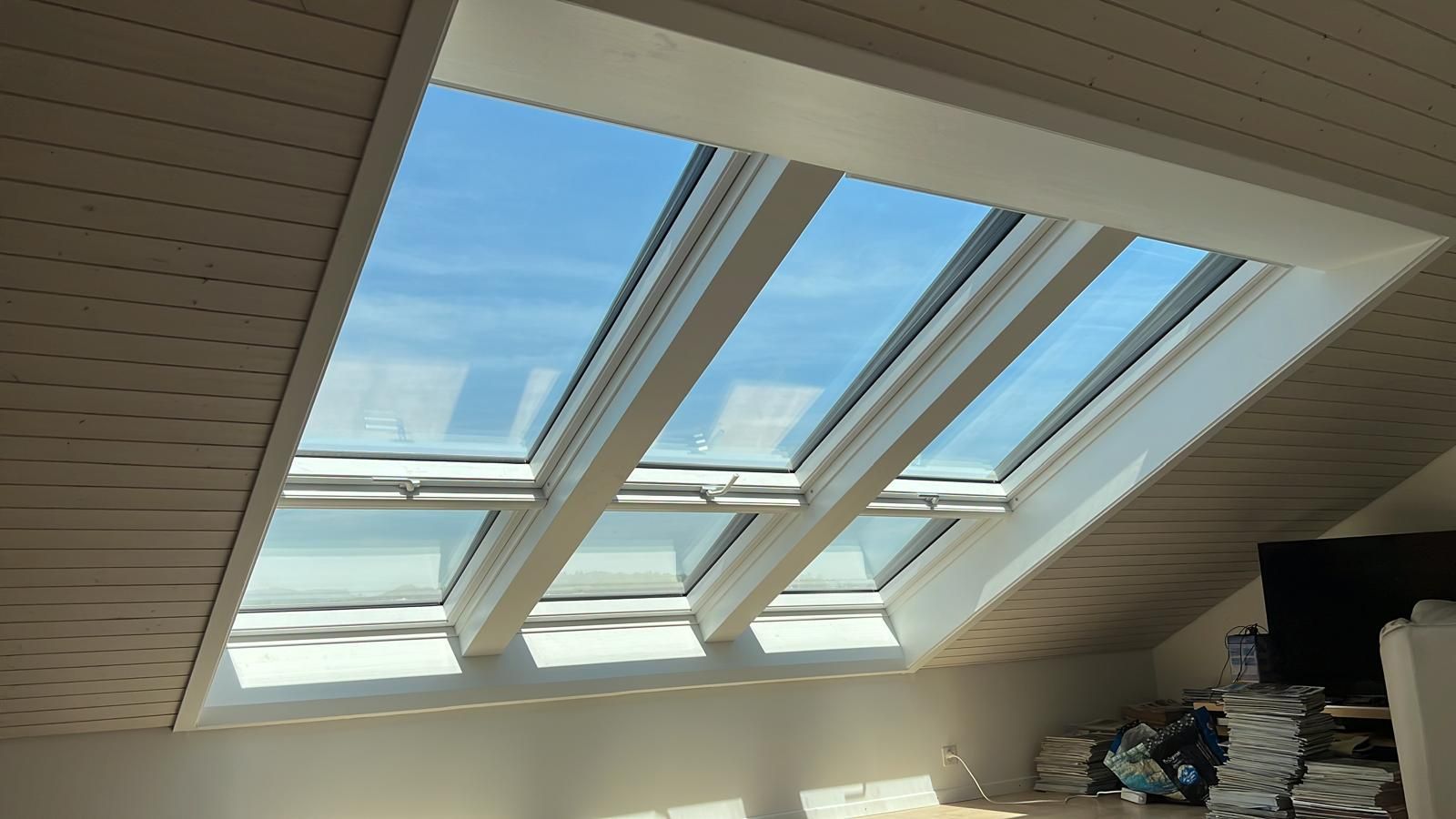 Drei schräg in einer hellen Decke angeordnete Oberlichter, weiß umrahmt, mit Blick auf einen blauen Himmel.