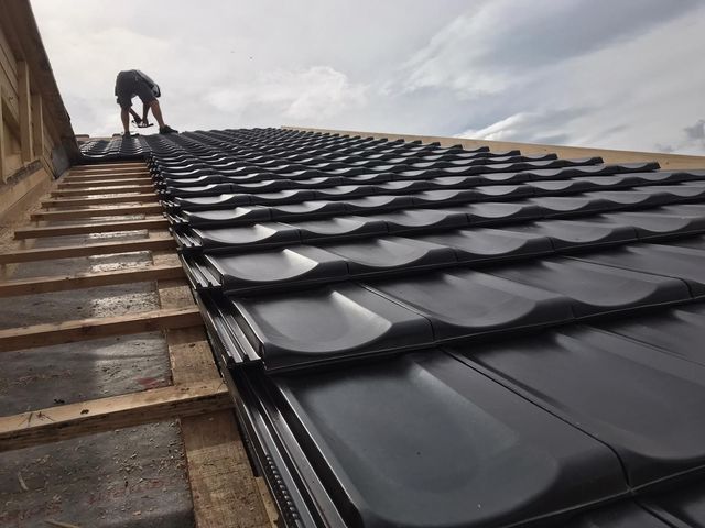 Dachdecker installiert dunkelgraue Metalldachziegel auf einer Holzkonstruktion vor einem bewölkten Himmel.