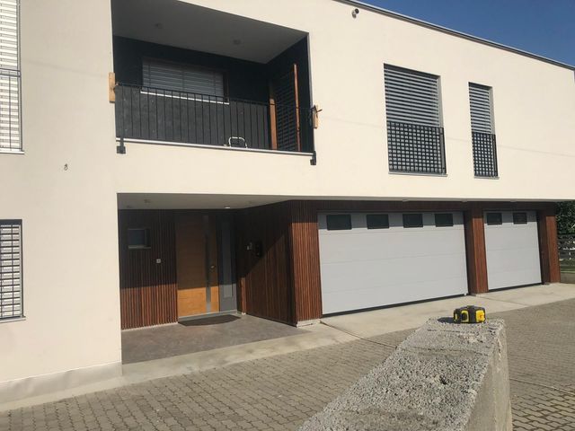 Modernes zweistöckiges Haus mit hellbeigen Wänden, dunkelbraunen Akzenten und grauen Garagentoren an einem sonnigen Tag.