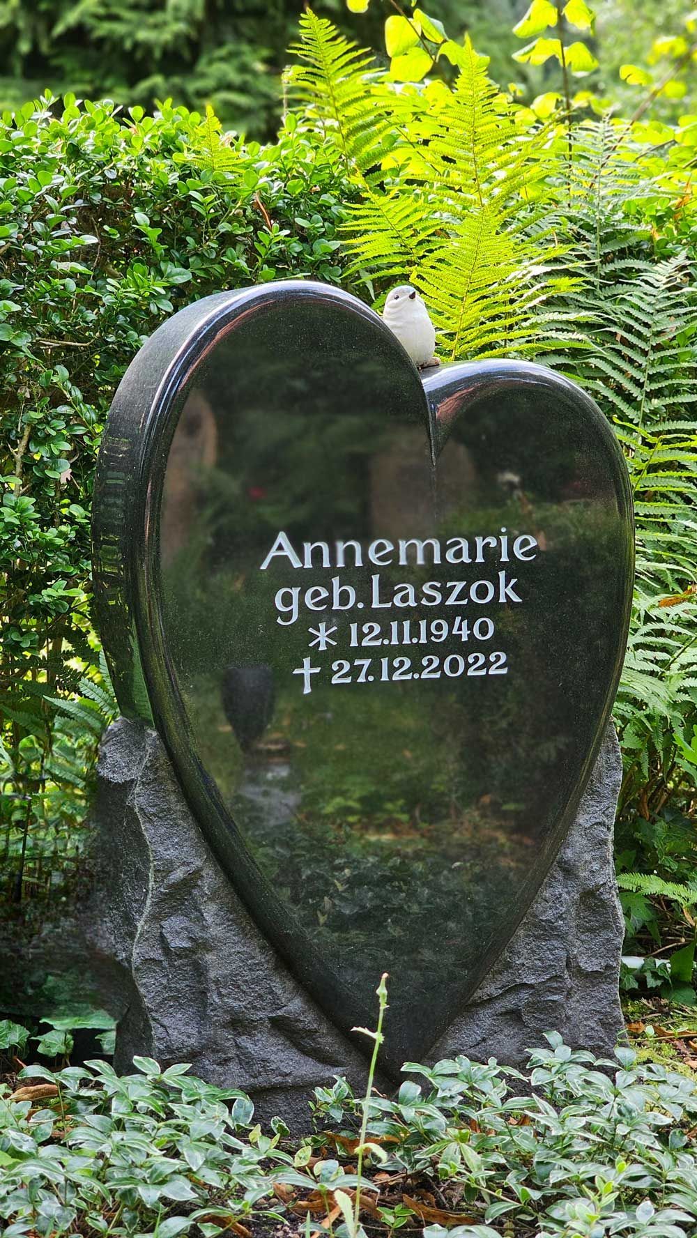 Herzförmiger Grabstein aus schwarzem Granit; Inschrift: Annemarie, geb. Laszok, * 12.11.1940, † 27.12.2022. Eingefasst in grünes Laubwerk.