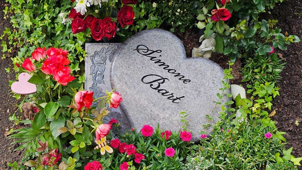 Herzförmiger Grabstein mit der Aufschrift „Simone Bart“, umgeben von roten Rosen und Grünpflanzen.
