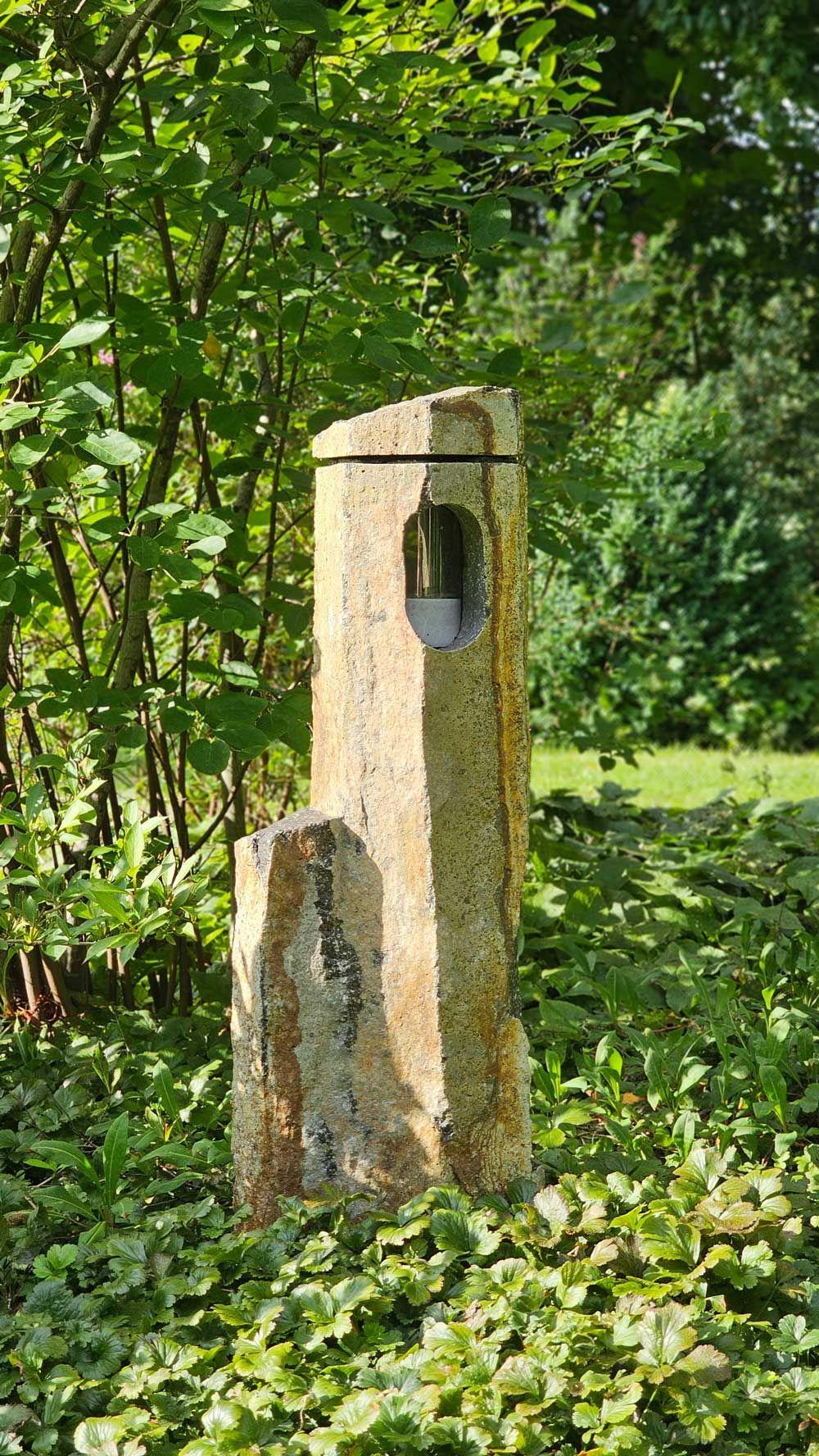 Eine hohe, verwitterte Steinskulptur mit einem kreisrunden Loch, umgeben von üppigem Grün.